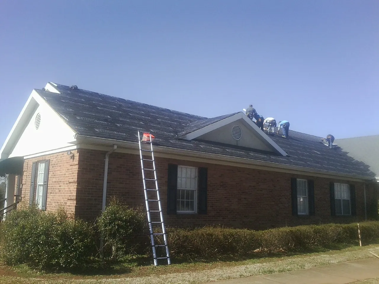 Crew tearing shingles off a brick ranch roof with a ladder set — Emergency Roof Repair work by our crew in White Pine