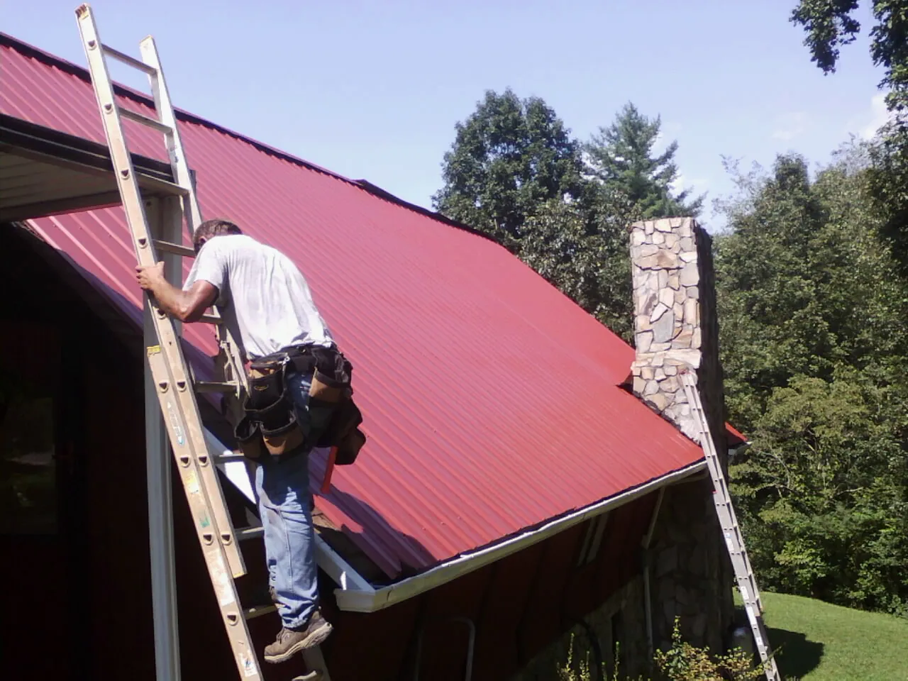 Roofer on a ladder beside a red metal roof and stone chimney — Soffit & Fascia Repair by Roofing Brothers in White Pine